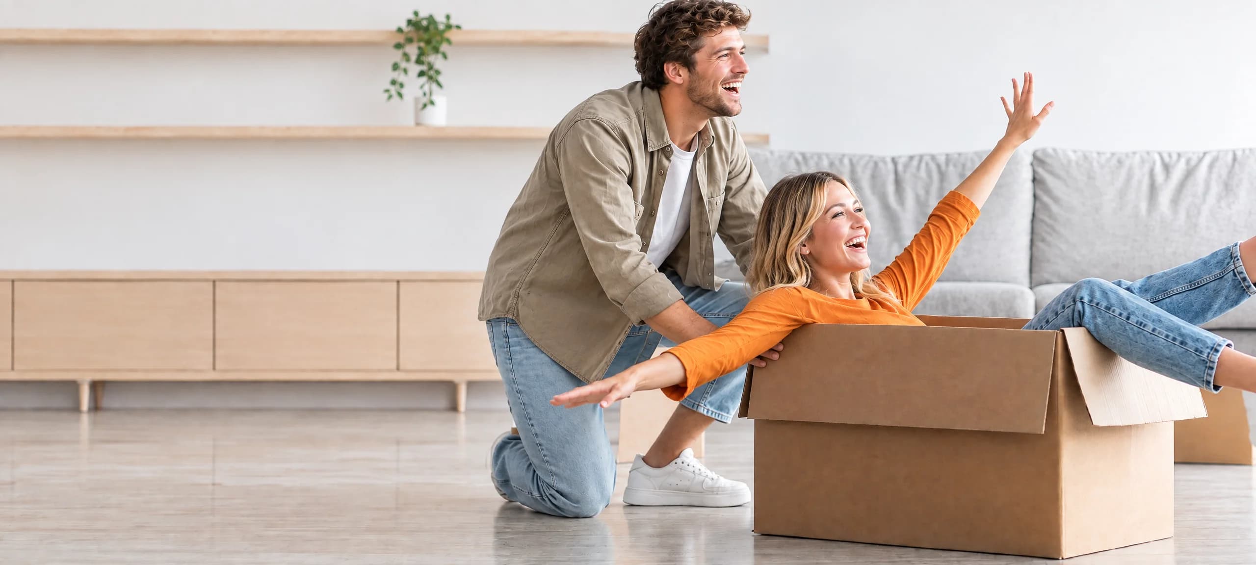 A couple laughing in their new living room, one pushing the other along in a moving box